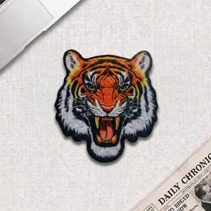 May include: Embroidered patch featuring a detailed tiger head with an open mouth, showcasing sharp teeth. The tiger has orange, black, and white fur, with yellow accents. The patch is likely for clothing or accessories.