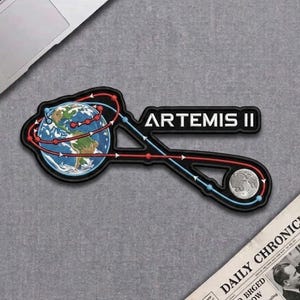 May include: Embroidered patch featuring the words "ARTEMIS II" in white block letters. The patch design includes a globe, a depiction of the moon, and a stylized orbital path in red and blue. The patch has a black border.