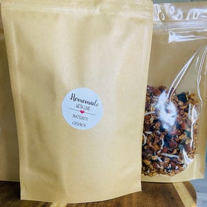 May include: Brown paper bags of granola with a clear plastic bag filled with granola next to it. The brown paper bag has a white sticker that says "Homemade with Love" and "Nature's Crunch". The granola appears to have nuts and coconut flakes.