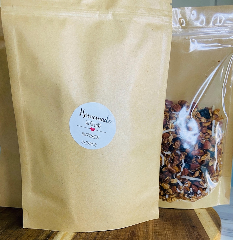 May include: Brown paper bags of granola with a clear plastic bag filled with granola next to it. The brown paper bag has a white sticker that says "Homemade with Love" and "Nature's Crunch". The granola appears to have nuts and coconut flakes.
