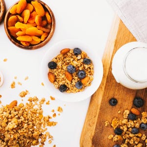 Könnte beinhalten: Draufsicht auf ein gesundes Frühstück. Schalen mit Cashewnüssen und getrockneten Aprikosen sind zusammen mit einer Schüssel mit Müsli mit Blaubeeren und Mandeln angeordnet. Ein Glas Milch und ein Holzbrett sind ebenfalls vorhanden.
