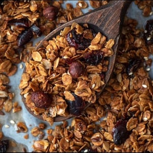May include: Close-up of homemade granola in a wooden spoon. The granola is a mix of golden-brown oats, nuts, and dried fruit, including dark red cranberries and hazelnuts. The spoon is filled with granola, with more granola scattered around.