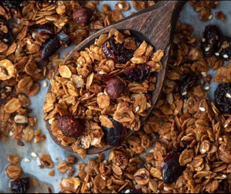May include: Close-up of homemade granola in a wooden spoon. The granola is a mix of golden-brown oats, nuts, and dried fruit, including dark red cranberries and hazelnuts. The spoon is filled with granola, with more granola scattered around.