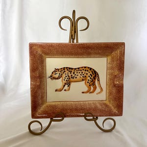 May include: A rectangular ceramic tray featuring a hand-painted leopard in the center, set against a cream-colored background. The tray has a wide, textured brown border and is displayed on a gold-toned metal stand with decorative scrollwork.