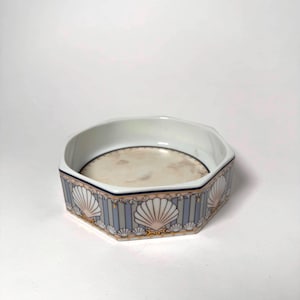 May include: A white ceramic hexagonal bowl featuring a decorative exterior pattern of light blue vertical stripes, gold-toned accents, and repeating white seashell motifs. The interior base of the bowl has a smooth, light beige marbled finish.