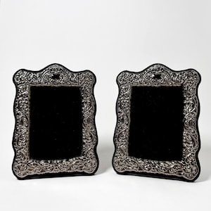 May include: Two rectangular picture frames with a metallic silver finish and intricate embossed scrollwork patterns. Each frame features a scalloped edge and a solid black rectangular center insert, providing a high-contrast display for photographs.