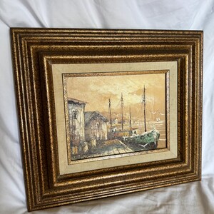 May include: A framed oil painting depicting a coastal harbor scene with a green boat docked near rustic buildings. The artwork features a warm, sepia-toned sky and is encased in a wide, ornate, textured gold-colored frame with a beige fabric mat.