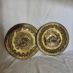 May include: Two circular brass decorative plates featuring a central embossed design of a basket filled with fruit. The plates have a wide, ornate border with repeating leaf-like patterns and a polished, reflective gold-toned metallic finish.