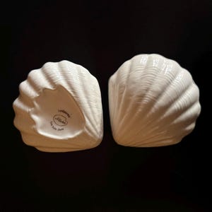 May include: Two white, hand-painted, fine bone china seashell-shaped dishes. The dishes have a ribbed texture and are set against a black background. The text "handpainted" and "fine bone china" is visible on the bottom of one dish.