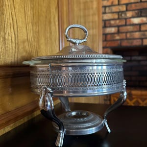 May include: Silver-plated chafing dish with a clear glass lid and decorative pierced metalwork. The dish has a handle on the lid and is supported by ornate legs. A classic piece for serving food.