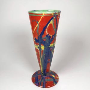 May include: A conical glass vase featuring a vibrant red base color with abstract splatters and streaks of blue, yellow, and green. The vase has a flared rim, a tapered body, and a circular footed base, all decorated with a multi-colored paint pattern.