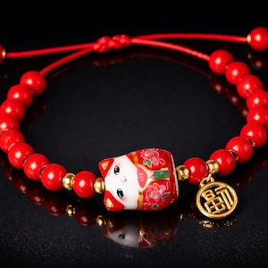 Peut inclure: Bracelet de perles rouges avec un charm en céramique Maneki-neko, des détails floraux et un charm doré. Le bracelet est monté sur un cordon rouge et est réglable.
