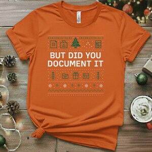 Può includere: T-shirt arancione con un design grafico verde e bianco. Il design include codice binario, alberi di Natale e la scritta "BUT DID YOU DOCUMENT IT". La maglietta è realizzata in materiale morbido e ha maniche corte.