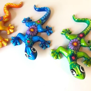May include: Three colorful, hand-painted ceramic gecko figurines. The geckos are in yellow, blue, and green, each with floral patterns and detailed eyes. The gecko's tails are curled.