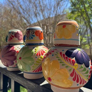 Pode incluir: Três copos de shot de tequila de cerâmica pintados à mão com tampas a condizer. Os copos de shot são redondos com desenhos florais em tons de rosa, verde, amarelo e azul. Os copos de shot estão sobre um corrimão de madeira.