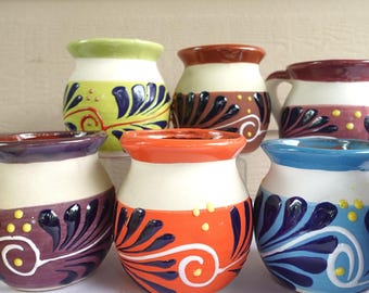 Könnte beinhalten: Eine Sammlung von sechs kleinen, handbemalten Keramikbechern. Jeder Becher hat ein einzigartiges Farbschema, darunter Grün, Orange, Lila und Blau, mit weißen und braunen Akzenten. Die Becher sind mit stilisierten Blumen- und Wirbelmustern verziert.
