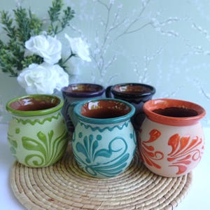 May include: A collection of five rounded ceramic mugs featuring hand-painted floral patterns in shades of lime green, teal, orange, purple, and black. Each mug has a smooth, curved body with a wide rim and a natural terracotta-colored interior.