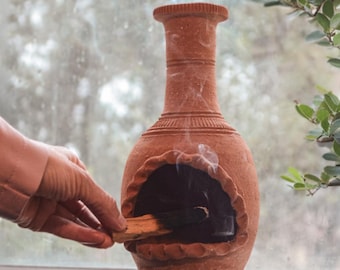 Pode incluir: Um queimador de incenso de terracota com corpo redondo, três pés pequenos e um pescoço alto. A fumaça sobe da abertura onde um pau de incenso está sendo segurado. O queimador é colocado em uma janela com fundo desfocado.