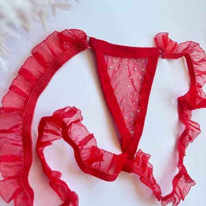 Può includere: Completo intimo rosso con perizoma con pannello in rete trasparente ornato da piccoli abbellimenti scintillanti. Il set include finiture a balze lungo i bordi e le spalline, creando un look delicato e femminile. Il tessuto appare leggero e semitrasparente.