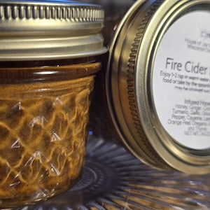 May include: Close-up of two small glass jars of Fire Cider Honey. The jars have gold-colored lids and are filled with a golden-brown liquid. The label on one jar lists ingredients: honey, ginger, turmeric, garlic, onion, black pepper, cayenne, lemon, orange peel, oregano, rosemary, and thyme.