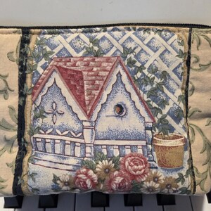 May include: A rectangular fabric pouch with a house and floral design. The center features a house with a red roof, blue walls, and a white trellis. The sides have a beige background with green floral patterns. The pouch is closed with a black zipper.
