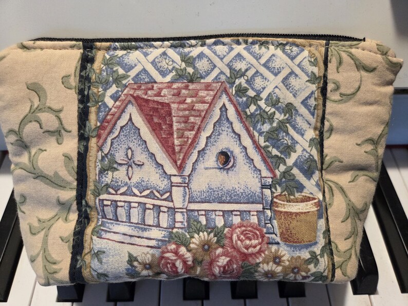 May include: A rectangular fabric pouch with a house and floral design. The center features a house with a red roof, blue walls, and a white trellis. The sides have a beige background with green floral patterns. The pouch is closed with a black zipper.