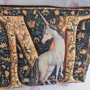 May include: A zippered pouch featuring a detailed illustration of a white unicorn with a long horn, set against a backdrop of the letter "M" and floral patterns in shades of gold, blue, and brown. The pouch has a dark brown zipper.