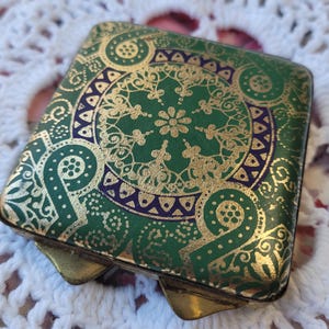May include: A vintage green and gold compact with a decorative floral pattern. The compact has a hinged lid and a brass frame.
