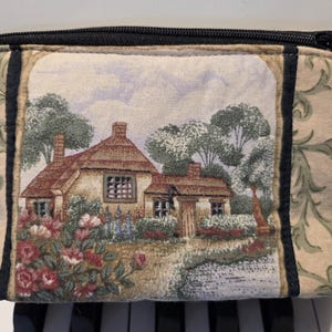 May include: A rectangular beige cosmetic bag with a black zipper. The bag features a tapestry design of a cottage with a thatched roof, surrounded by trees and flowers. The sides have a green and beige floral pattern.