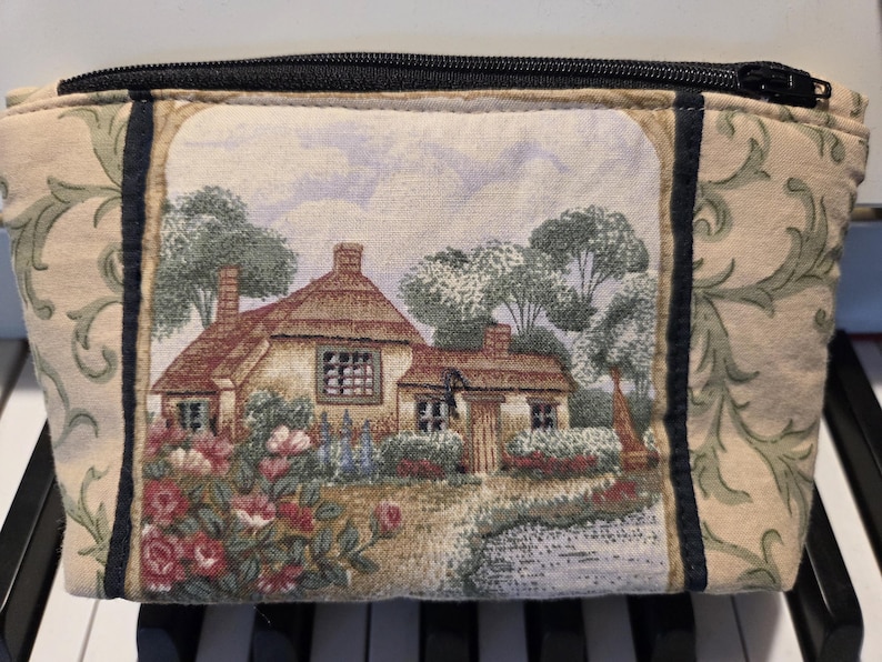 May include: A rectangular beige cosmetic bag with a black zipper. The bag features a tapestry design of a cottage with a thatched roof, surrounded by trees and flowers. The sides have a green and beige floral pattern.