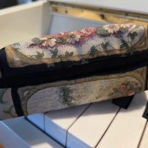 May include: A rectangular floral print fabric pouch with a black zipper. The pouch features a pattern of roses, daisies, and foliage in shades of pink, cream, and green. The pouch is resting on a white surface.