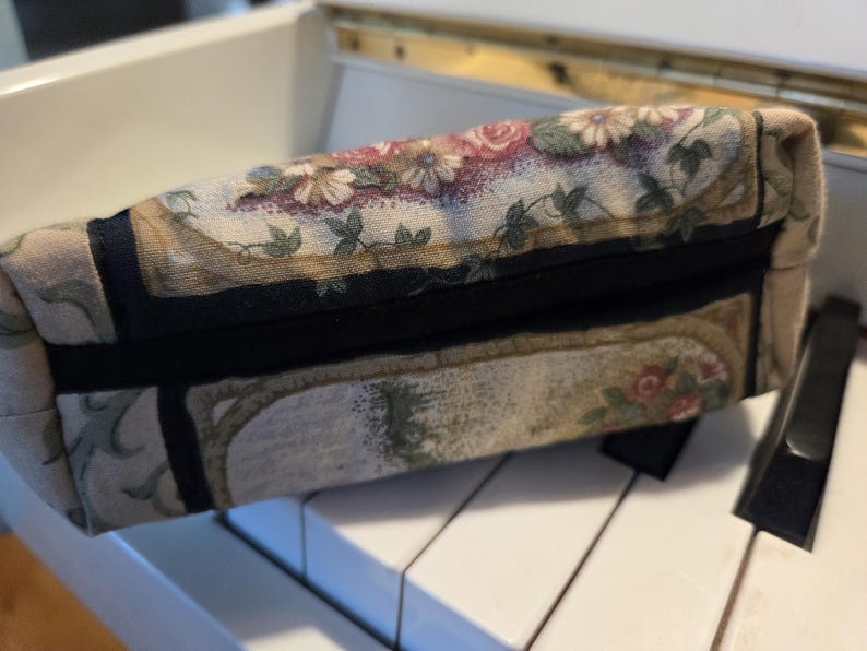 May include: A rectangular floral print fabric pouch with a black zipper. The pouch features a pattern of roses, daisies, and foliage in shades of pink, cream, and green. The pouch is resting on a white surface.