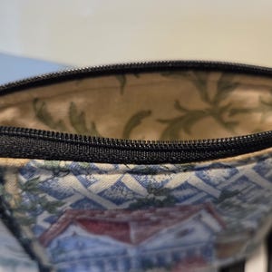 May include: Close-up of a small, open zippered pouch. The exterior fabric features a blue and white pattern with a house design. The interior lining is a light beige with green leaf patterns. The zipper is black.