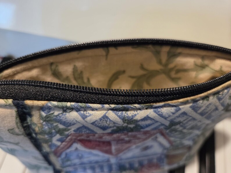 May include: Close-up of a small, open zippered pouch. The exterior fabric features a blue and white pattern with a house design. The interior lining is a light beige with green leaf patterns. The zipper is black.