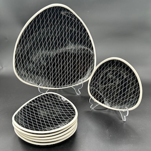 May include: A set of black triangular plates with a white grid pattern and white rims. The plates are displayed on clear stands against a dark background. The set includes large serving plates and smaller individual plates.