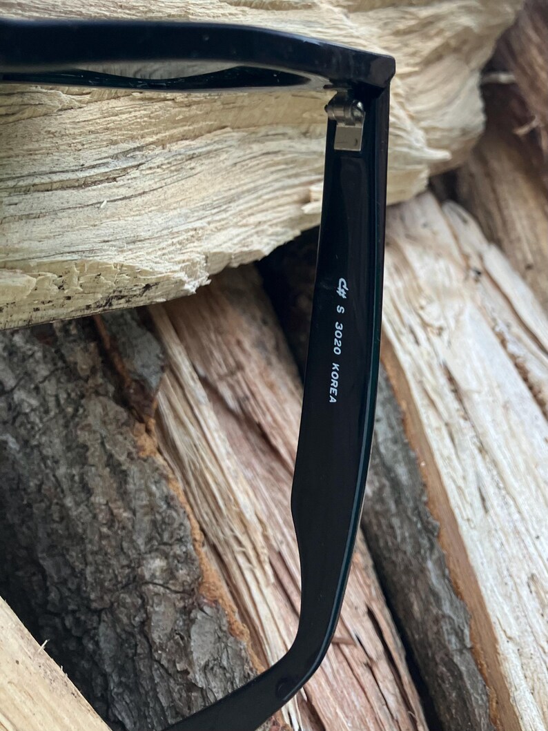 Puede incluir: Primer plano de gafas de sol negras con el texto "cff S 3020 KOREA" impreso en la patilla. Las gafas de sol est&aacute;n sobre un fondo de madera marr&oacute;n texturizada.