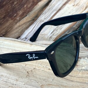 Puede incluir: Gafas de sol Ray-Ban negras con lentes verde oscuro. Las gafas tienen un dise&ntilde;o de montura angular cl&aacute;sico. El logotipo de Ray-Ban es visible en blanco en la patilla izquierda. Las gafas descansan sobre un trozo de madera.