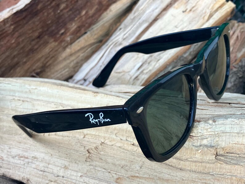 Puede incluir: Gafas de sol Ray-Ban negras con lentes verde oscuro. Las gafas tienen un dise&ntilde;o de montura angular cl&aacute;sico. El logotipo de Ray-Ban es visible en blanco en la patilla izquierda. Las gafas descansan sobre un trozo de madera.