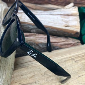 Puede incluir: Gafas de sol negras con el logotipo de Ray-Ban en escritura blanca en la patilla. Las gafas de sol descansan sobre una superficie de madera. Las lentes son oscuras y las monturas son de color negro brillante.