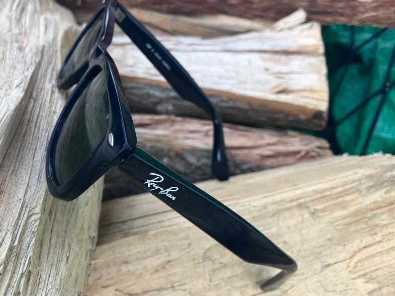 Puede incluir: Gafas de sol negras con el logotipo de Ray-Ban en escritura blanca en la patilla. Las gafas de sol descansan sobre una superficie de madera. Las lentes son oscuras y las monturas son de color negro brillante.