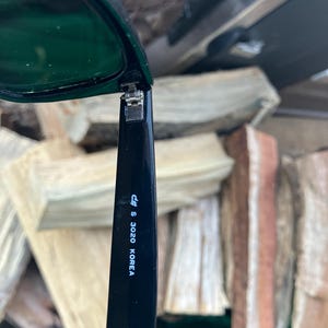 Puede incluir: Gafas de sol negras con lentes verde oscuro. La patilla de las gafas tiene el texto "c# S 3020 KOREA" impreso en blanco. Las gafas est&aacute;n abiertas, revelando la bisagra.
