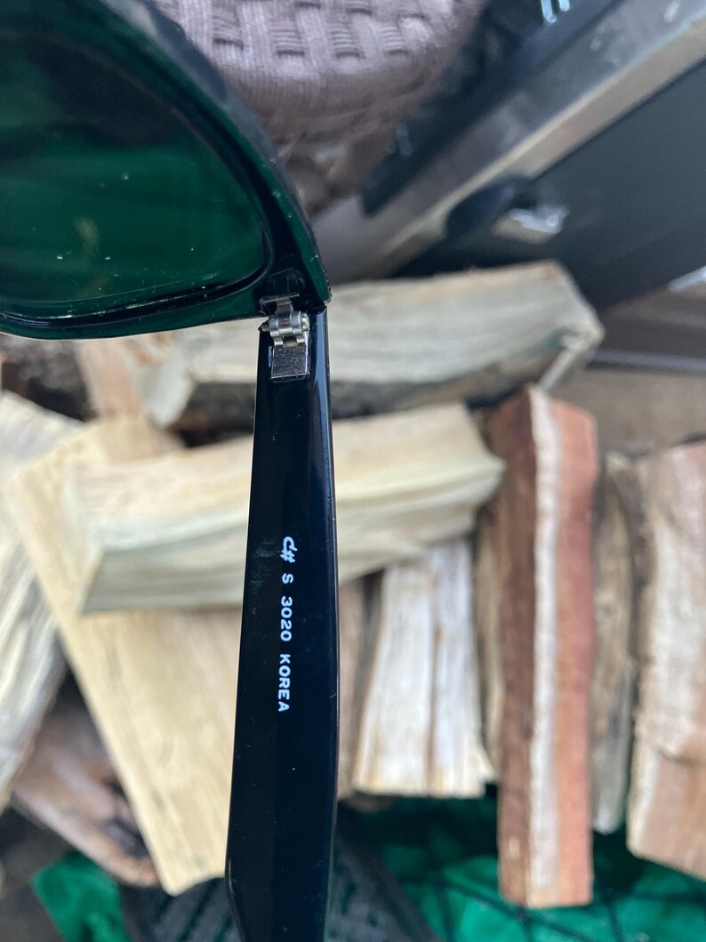 Puede incluir: Gafas de sol negras con lentes verde oscuro. La patilla de las gafas tiene el texto "c# S 3020 KOREA" impreso en blanco. Las gafas est&aacute;n abiertas, revelando la bisagra.