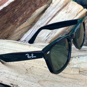 Puede incluir: Gafas de sol Ray-Ban negras con lentes oscuras. El logotipo de Ray-Ban es visible en la patilla izquierda. Las gafas de sol est&aacute;n sobre un trozo de madera. Dise&ntilde;o cl&aacute;sico y atemporal.