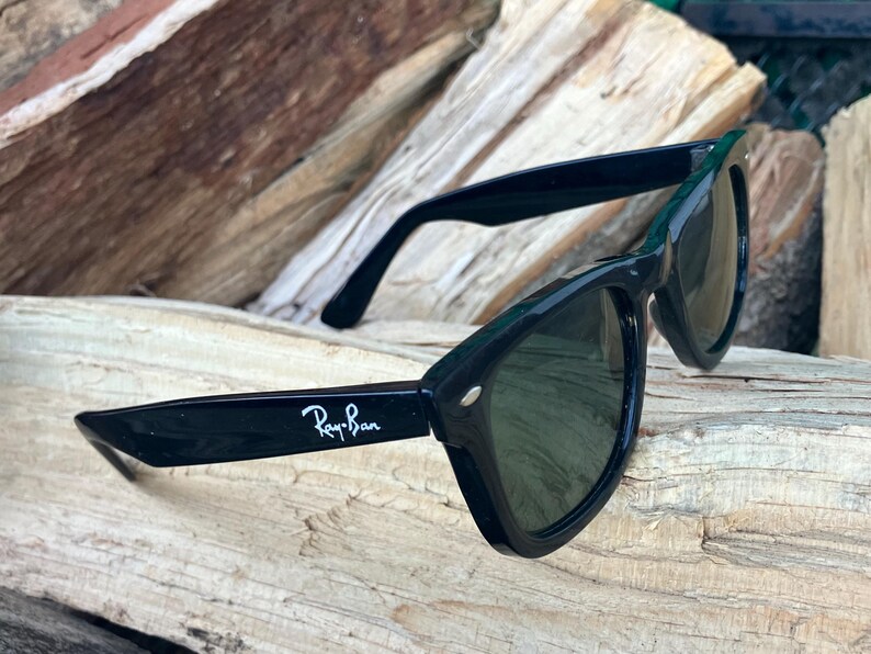 Puede incluir: Gafas de sol Ray-Ban negras con lentes oscuras. El logotipo de Ray-Ban es visible en la patilla izquierda. Las gafas de sol est&aacute;n sobre un trozo de madera. Dise&ntilde;o cl&aacute;sico y atemporal.