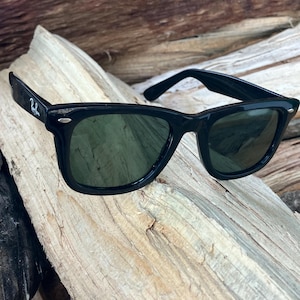 Puede incluir: Gafas de sol negras con lentes verde oscuro. Las gafas tienen una montura rectangular cl&aacute;sica. El logotipo de Ray-Ban es visible en la patilla izquierda. Las gafas descansan sobre un trozo de madera envejecida.