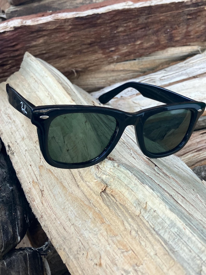 Puede incluir: Gafas de sol negras con lentes verde oscuro. Las gafas tienen una montura rectangular cl&aacute;sica. El logotipo de Ray-Ban es visible en la patilla izquierda. Las gafas descansan sobre un trozo de madera envejecida.