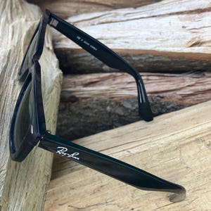 Puede incluir: Gafas de sol negras con el logotipo de Ray-Ban en la patilla. Las gafas de sol est&aacute;n sobre una superficie de madera. Las lentes son oscuras y las monturas son de color negro brillante. Las gafas de sol est&aacute;n en &aacute;ngulo, mostrando los detalles de la montura.