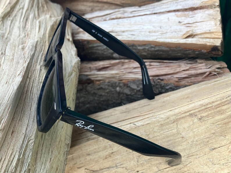 Puede incluir: Gafas de sol negras con el logotipo de Ray-Ban en la patilla. Las gafas de sol est&aacute;n sobre una superficie de madera. Las lentes son oscuras y las monturas son de color negro brillante. Las gafas de sol est&aacute;n en &aacute;ngulo, mostrando los detalles de la montura.