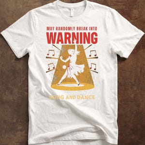 Puede incluir: Camiseta blanca con un diseño gráfico. El diseño presenta el texto "MAY RANDOMLY BREAK INTO WARNING SONG AND DANCE" en rojo y amarillo. Una silueta de un bailarín está en un foco, rodeada de notas musicales.