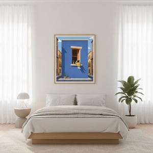 May include: A framed art print depicts a person in a yellow outfit sitting in a window, overlooking a street scene with buildings. The artwork has a blue wall and a wooden frame. The room has a bed, a side table with a lamp, and a potted plant.